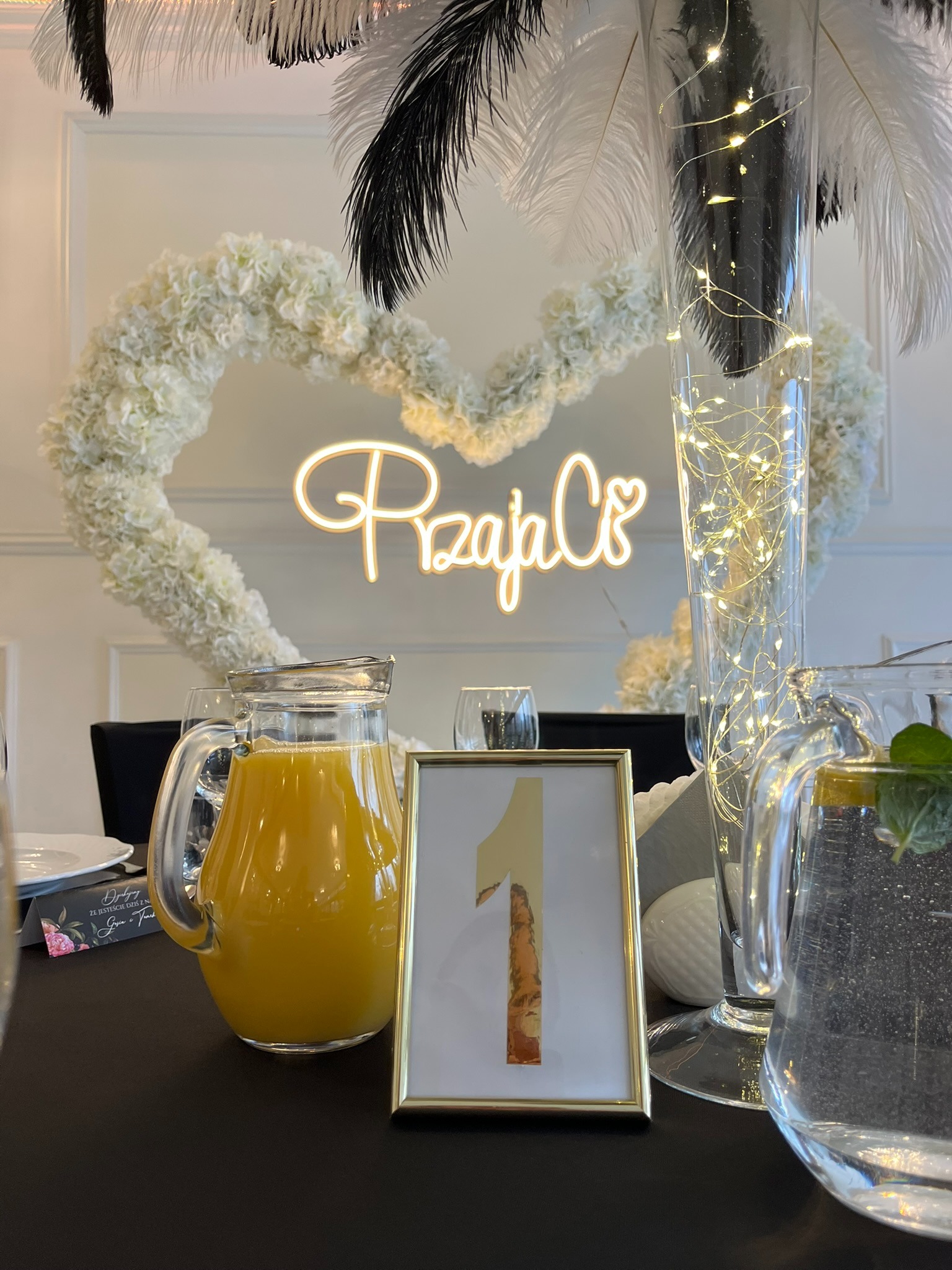 Dekoracja stołu z numerem '1' w złotej ramce, dzbanki z napojami, kwiatowe serce i pióra. Elegancki wystrój na przyjęcie.