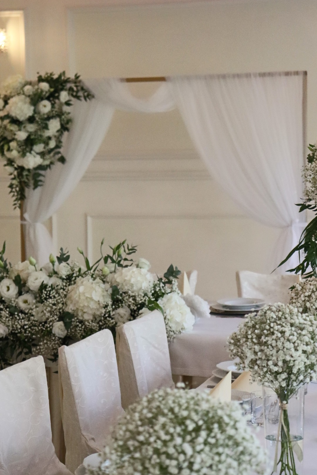 Elegancko udekorowany stół weselny z białymi kwiatami, subtelnymi dekoracjami i nakryciami, tworzący uroczystą atmosferę. Widoczne krzesła z białymi pokrowcami.