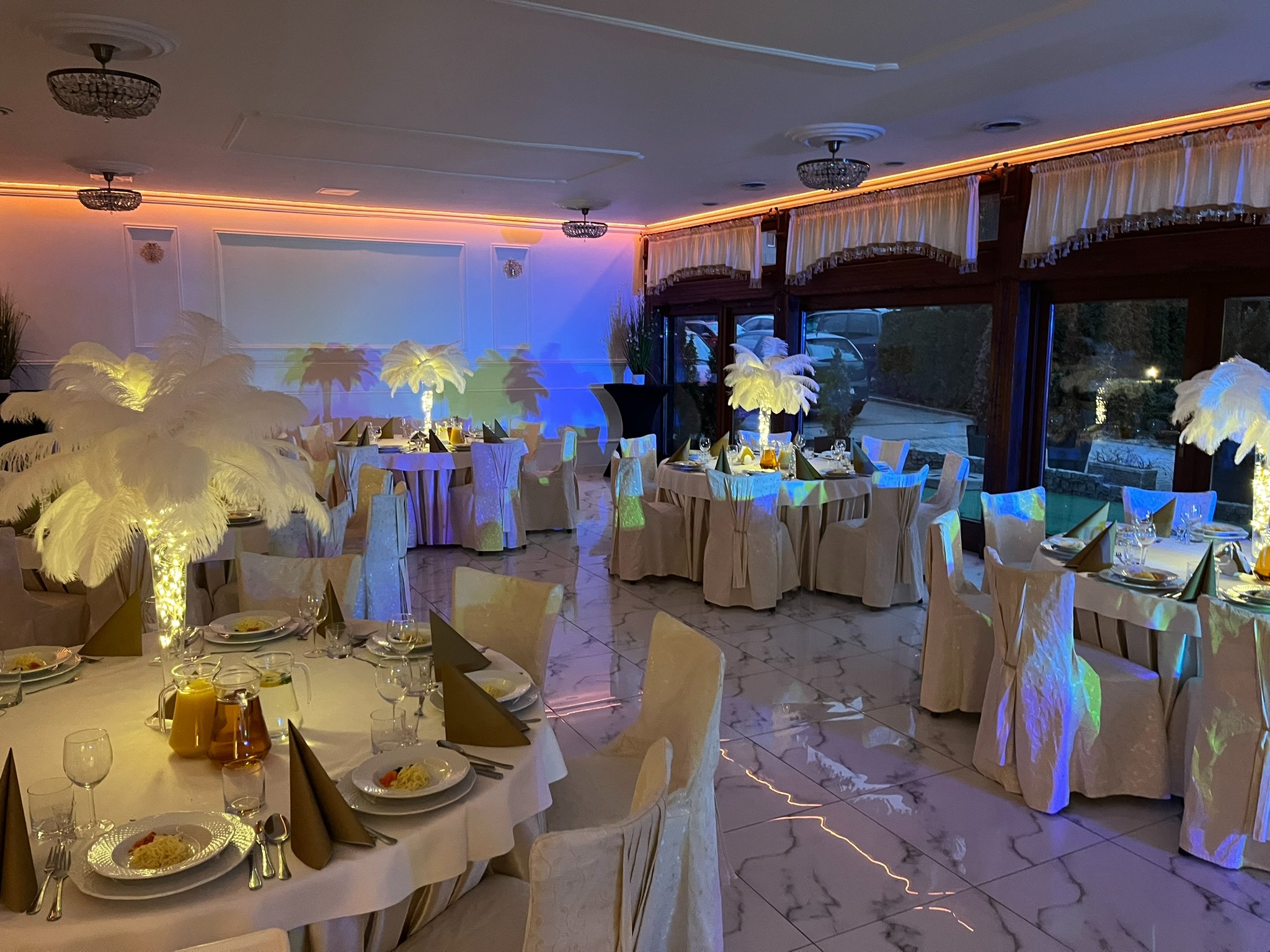 Elegancka sala bankietowa z okrągłymi stołami, białymi obrusami, piórowymi dekoracjami i subtelnym oświetleniem, gotowa na przyjęcie gości. Marmurowa podłoga.