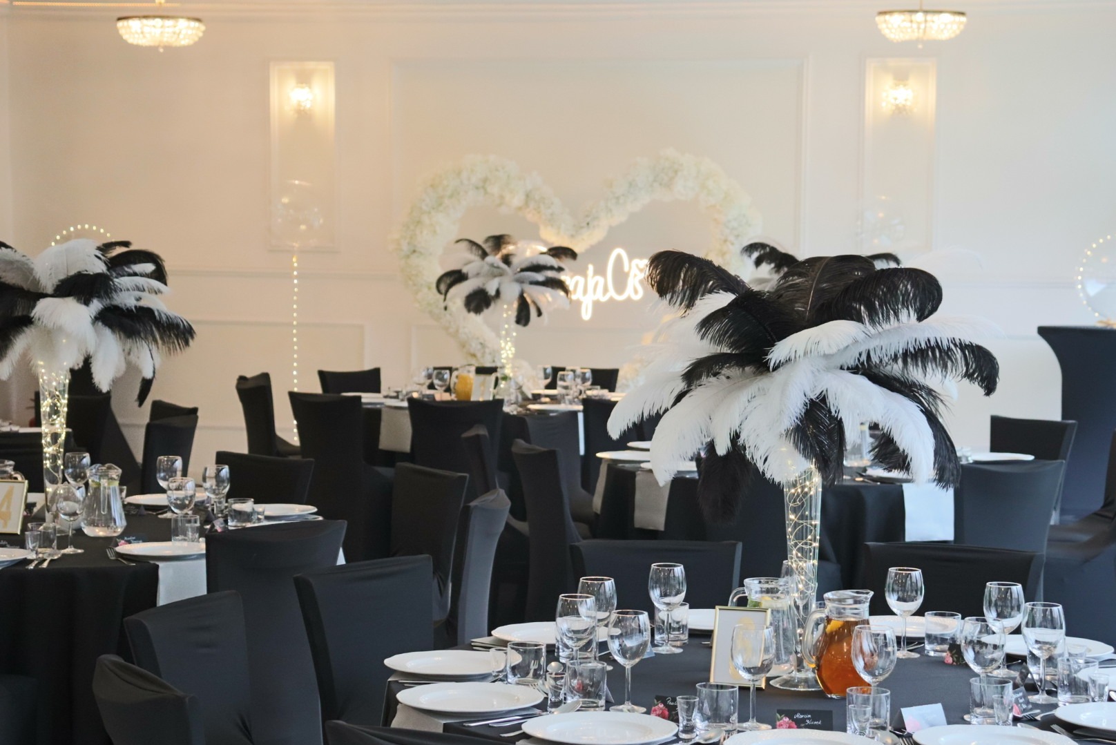 Elegancko nakryte stoły z czarnymi obrusami i białymi piórami w wazonach, dekoracja w kształcie serca z kwiatów i neonowy napis w tle, sala bankietowa.