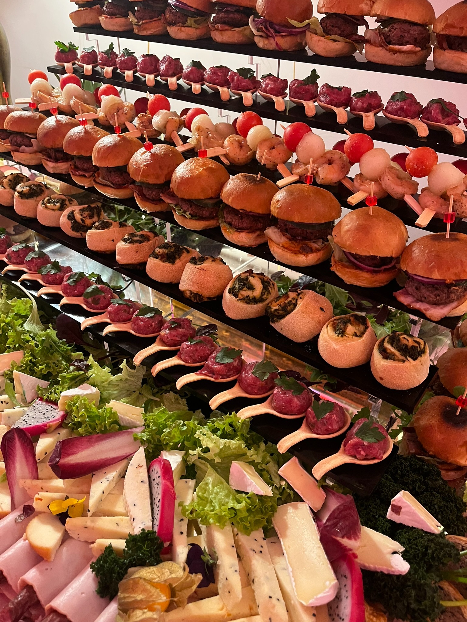 Elegancki stół cateringowy z mini burgerami, tartaletkami i przekąskami na łyżeczkach, uzupełniony deską serów i wędlin z egzotycznymi owocami.