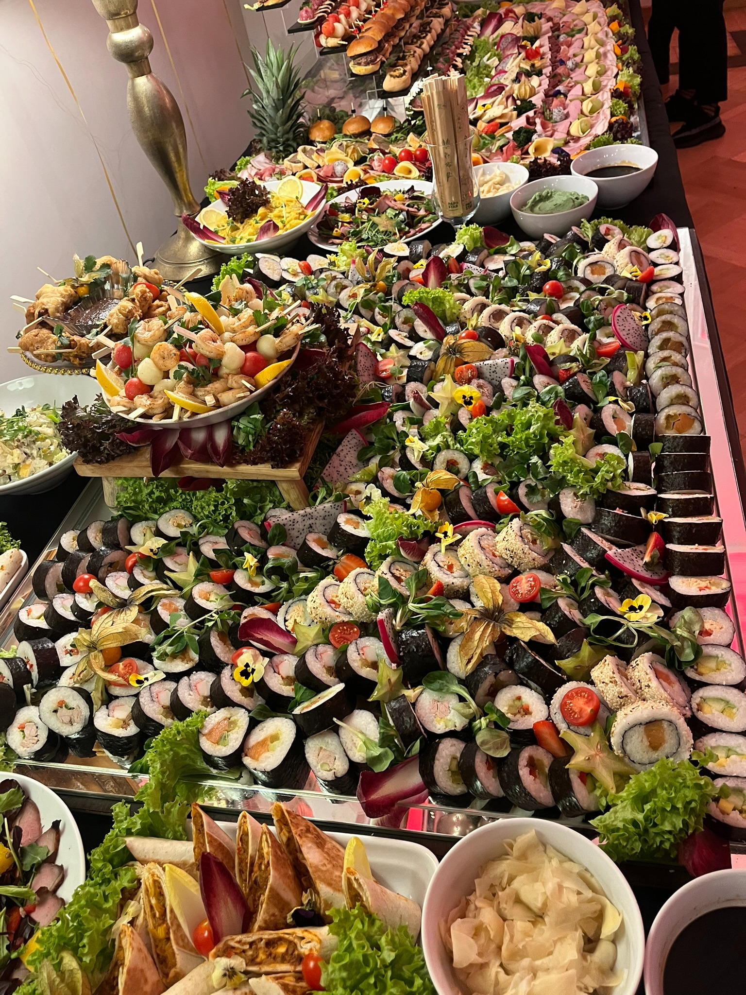 Bogato zastawiony stół cateringowy z sushi, przekąskami, sałatkami i owocami, udekorowany zielenią i kwiatami, tworzący elegancką kompozycję.