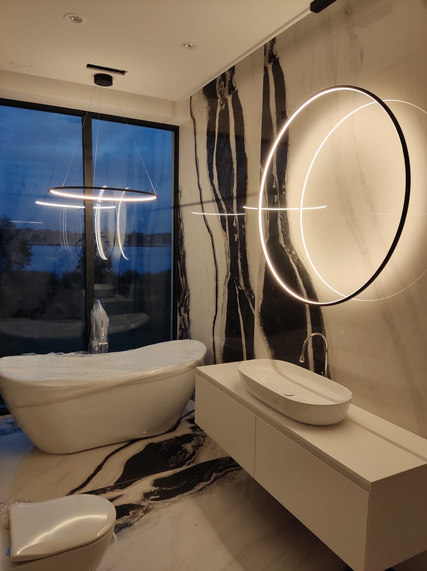 Nowoczesna łazienka z marmurowymi ścianami, owalną wanną w folii ochronnej i okrągłą lampą LED. Minimalistyczny design z widokiem na jezioro.