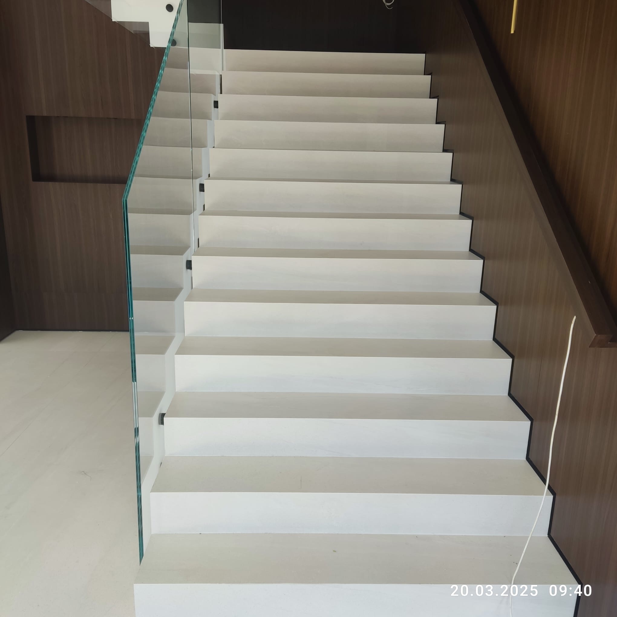 Nowoczesne, białe schody z szklaną balustradą i drewnianą okładziną ścienną. Minimalistyczny design, jasne wnętrze. Widok z dołu na górę.