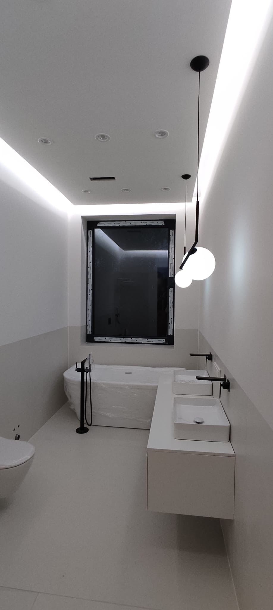 Nowoczesna łazienka z wanną owiniętą folią ochronną, podwieszanymi umywalkami i czarną armaturą. Oświetlenie LED i lampy wiszące dodają elegancji.