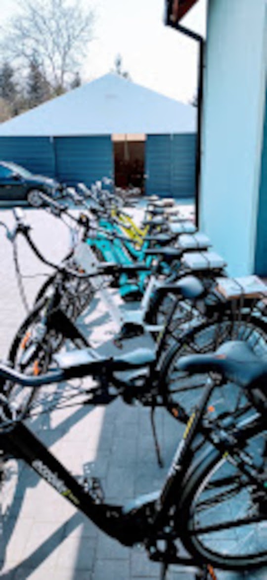 Rząd rowerów, w tym elektrycznych, zaparkowanych przed budynkiem z otwartymi drzwiami. Widoczne detale rowerów i cień rzucany na chodnik.