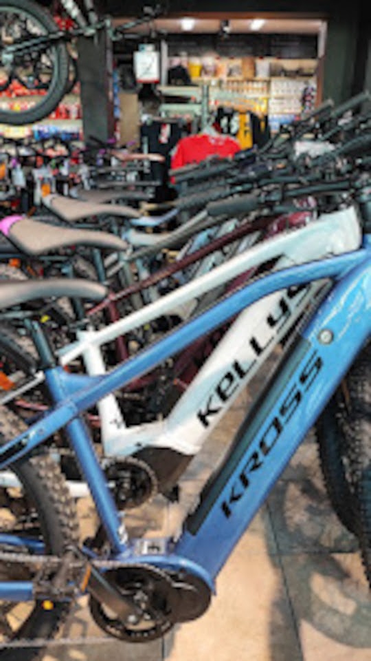 Rząd rowerów Kross i Kellys w sklepie, z niebieskim rowerem elektrycznym na pierwszym planie. Widoczne detale: opony, ramy, napisy. Tło z elementami wyposażenia sklepu.