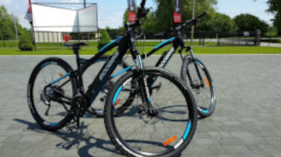 Dwa czarno-niebieskie rowery górskie na tle parku z zielenią i białym billboardem. Rowery ustawione obok siebie, z widocznymi detalami opon i ramy.