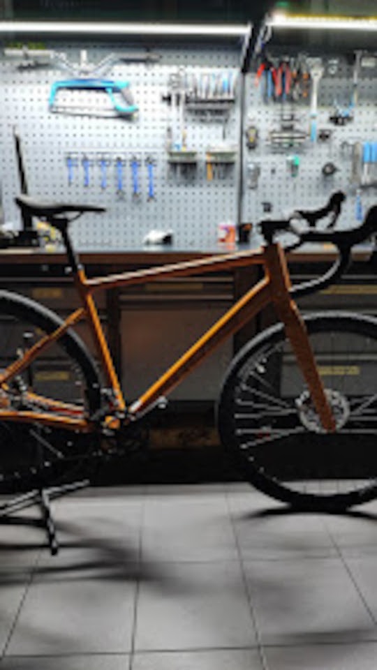 Rower gravelowy na stojaku w warsztacie, narzędzia na ścianie w tle. Pomarańczowa rama, czarne koła i siodełko. Drobne detale widoczne na tle warsztatowym.