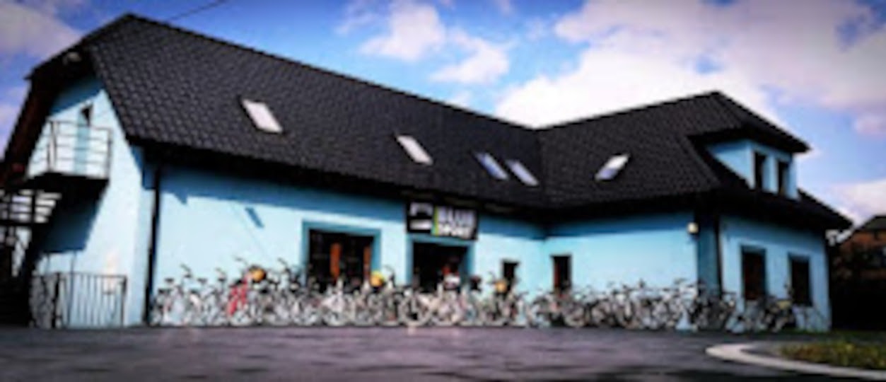 Błękitny budynek wypożyczalni rowerów z czarnym dachem i rzędem rowerów przed wejściem, pod błękitnym niebem z chmurami.