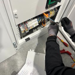 ALEKSANDER RUSAKOWICZ - Elektryk w trakcie pracy nad rozdzielnicą elektryczną, trzymający schemat. Widoczne narzędzia i elementy instalacji. Biała obudowa rozdzielnicy.