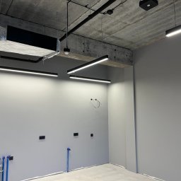 ALEKSANDER RUSAKOWICZ - Wnętrze biurowe z surowym betonowym sufitem i liniowym oświetleniem LED. Widoczne instalacje wentylacyjne i przygotowane punkty elektryczne na ścianie.