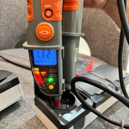 ALEKSANDER RUSAKOWICZ - Miernik napięcia w gnieździe, wyświetlający wartość 231V, z widocznymi przyciskami i diodami LED, na tle szarej sofy i kabli.