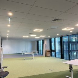 ALEKSANDER RUSAKOWICZ - Nowoczesne biuro z kwadratowymi lampami sufitowymi, jasną wykładziną i białymi biurkami. Widoczne okna z żaluzjami i elementy wentylacji.