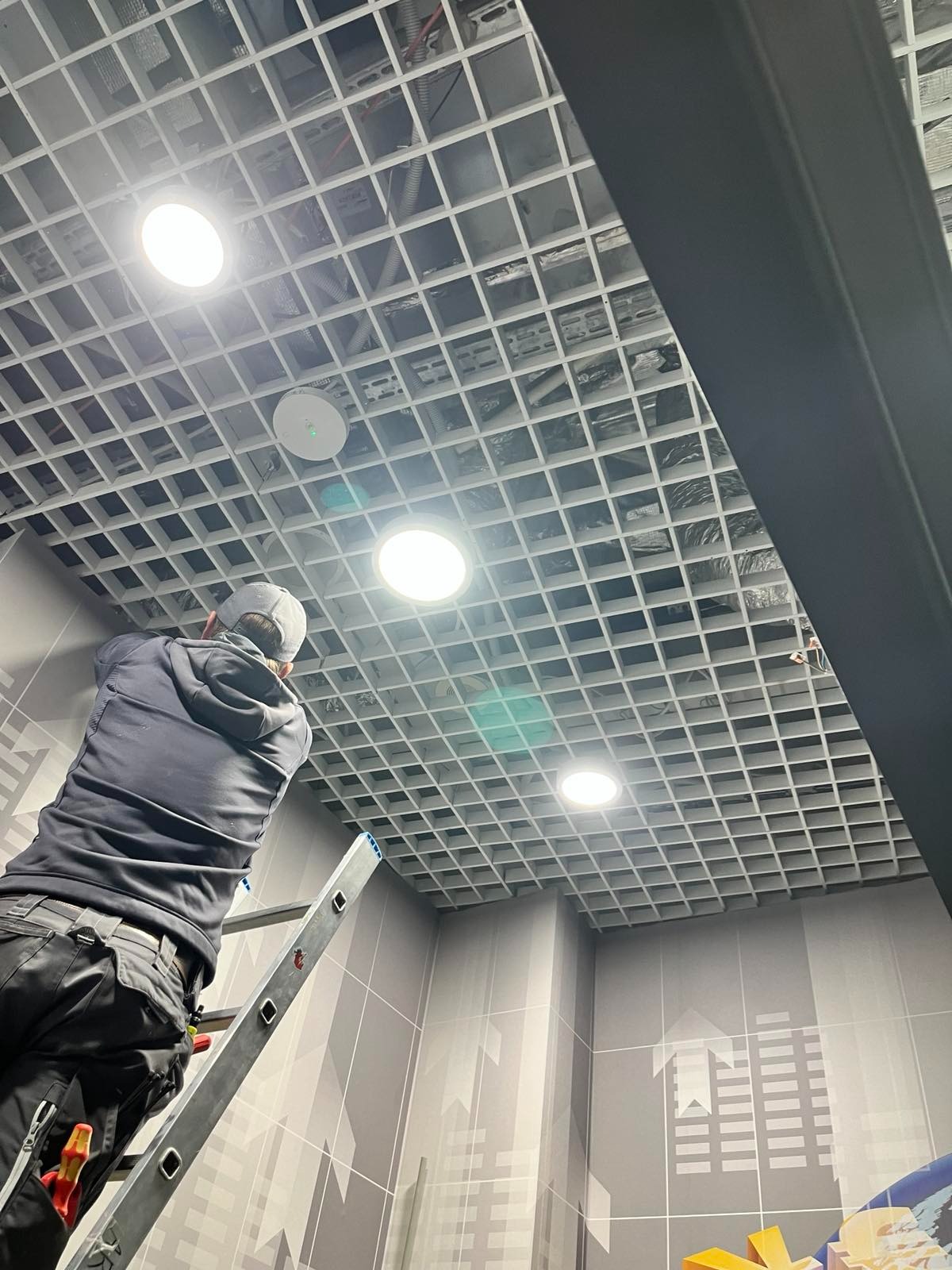 Pracownik na drabinie montuje oświetlenie w rastrowym suficie podwieszanym. Widoczne narzędzia i elementy instalacji. Ściany z motywem graficznym.