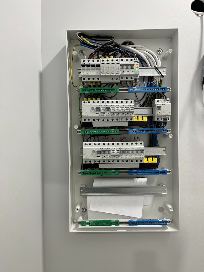 Rozdzielnica elektryczna w białej obudowie, widoczne zabezpieczenia, przewody i listwy zaciskowe. Nowoczesna instalacja w minimalistycznym wnętrzu.