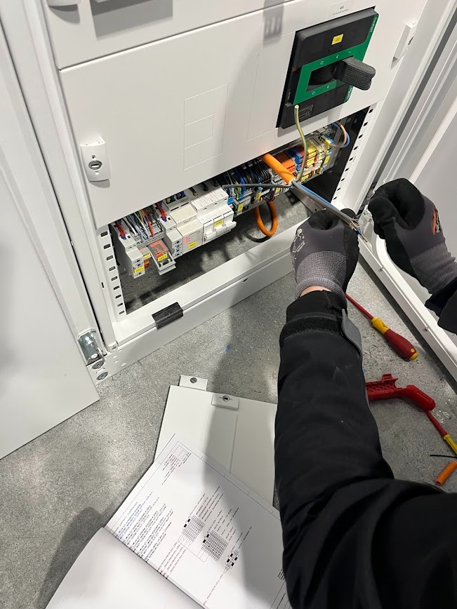 Elektryk w trakcie pracy nad rozdzielnicą elektryczną, trzymający schemat. Widoczne narzędzia i elementy instalacji. Biała obudowa rozdzielnicy.