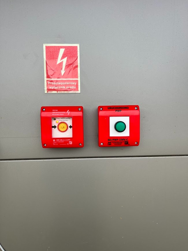 Dwa czerwone przyciski alarmowe (żółty i zielony) oraz naklejka ostrzegawcza z symbolem błyskawicy na szarej powierzchni. Przeciwpożarowy wyłącznik prądu.