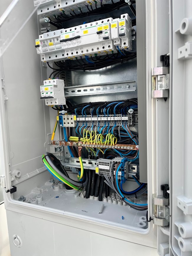 Wnętrze skrzynki elektrycznej z zamontowanymi bezpiecznikami, listwami zaciskowymi i okablowaniem w kolorach: niebieskim, czarnym, żółto-zielonym. Widoczne ślady montażu.