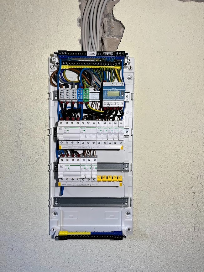 Rozdzielnica elektryczna z przewodami w ścianie, widok z przodu. Elementy zabezpieczeń, kable w różnych kolorach. Biała ściana z widocznymi śladami po instalacji.
