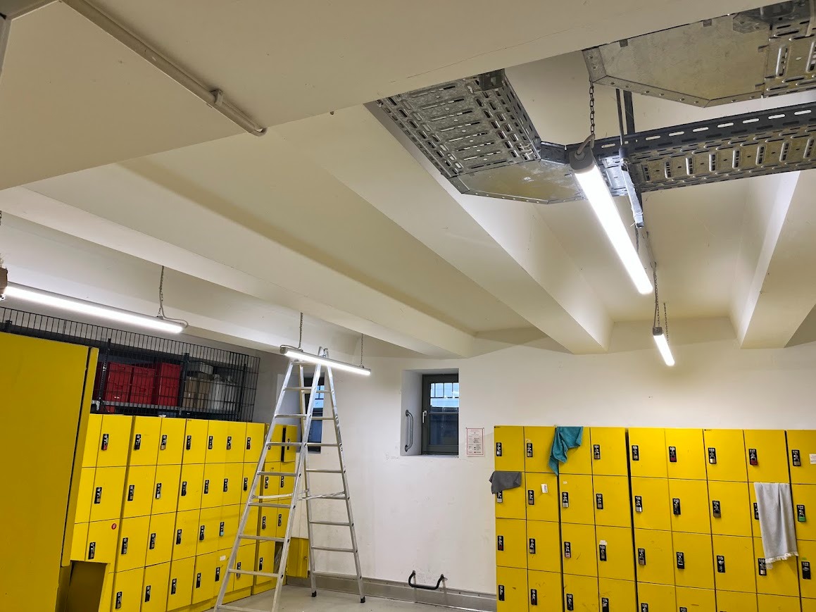 Wnętrze szatni z żółtymi szafkami, oświetlone wiszącymi lampami LED i metalowymi korytkami kablowymi pod sufitem. Aluminiowa drabina oparta o ścianę.