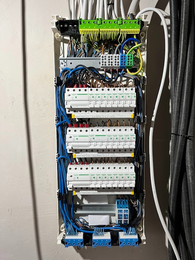 Rozdzielnica elektryczna Schneider z zabezpieczeniami, okablowaniem w kolorze niebieskim i czarnym, zamontowana na jasnej ścianie. Widoczne przewody w peszlu.