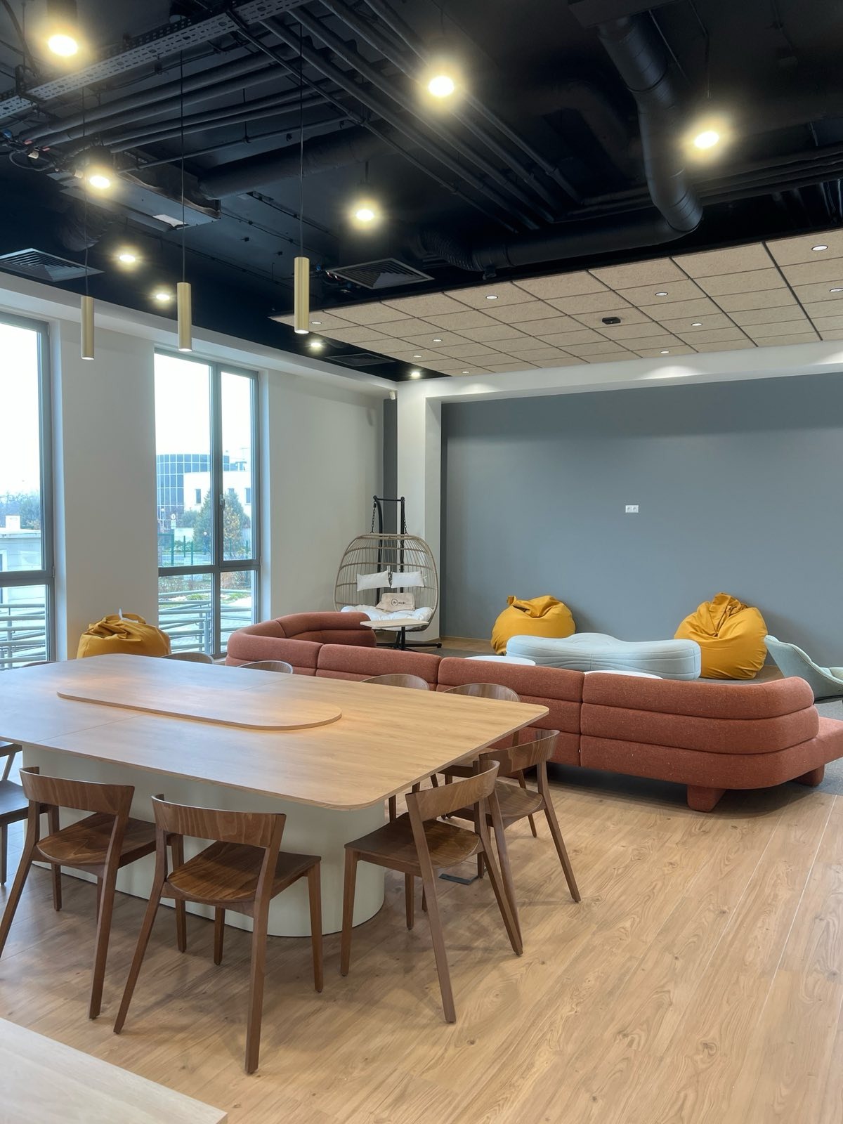 Nowoczesne biuro z widocznymi instalacjami pod sufitem, oświetleniem punktowym i wiszącymi lampami. Przestrzeń relaksu z sofą i pufami, oraz stół konferencyjny z krzesłami.