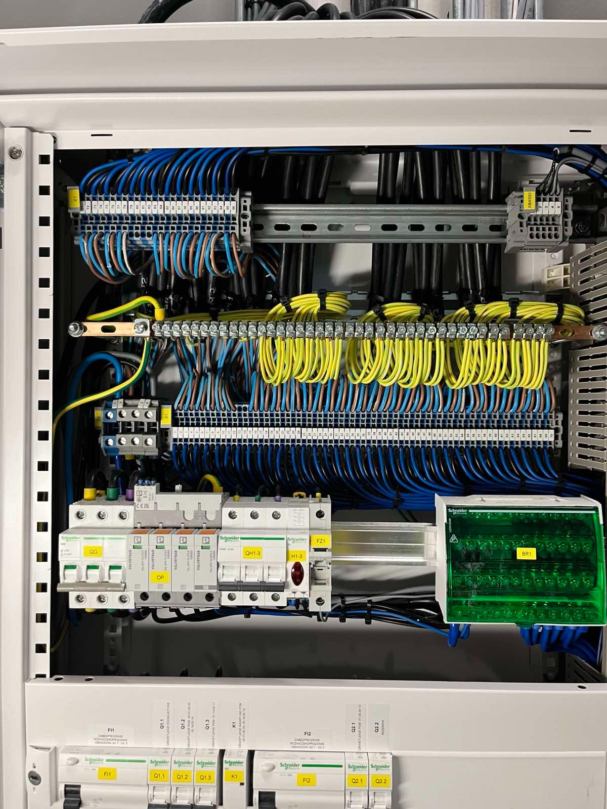 Rozdzielnica elektryczna z precyzyjnie ułożonymi przewodami w kolorach niebieskim, żółtym i czarnym. Widoczne bezpieczniki i oznaczenia. Estetyczne wykonanie.
