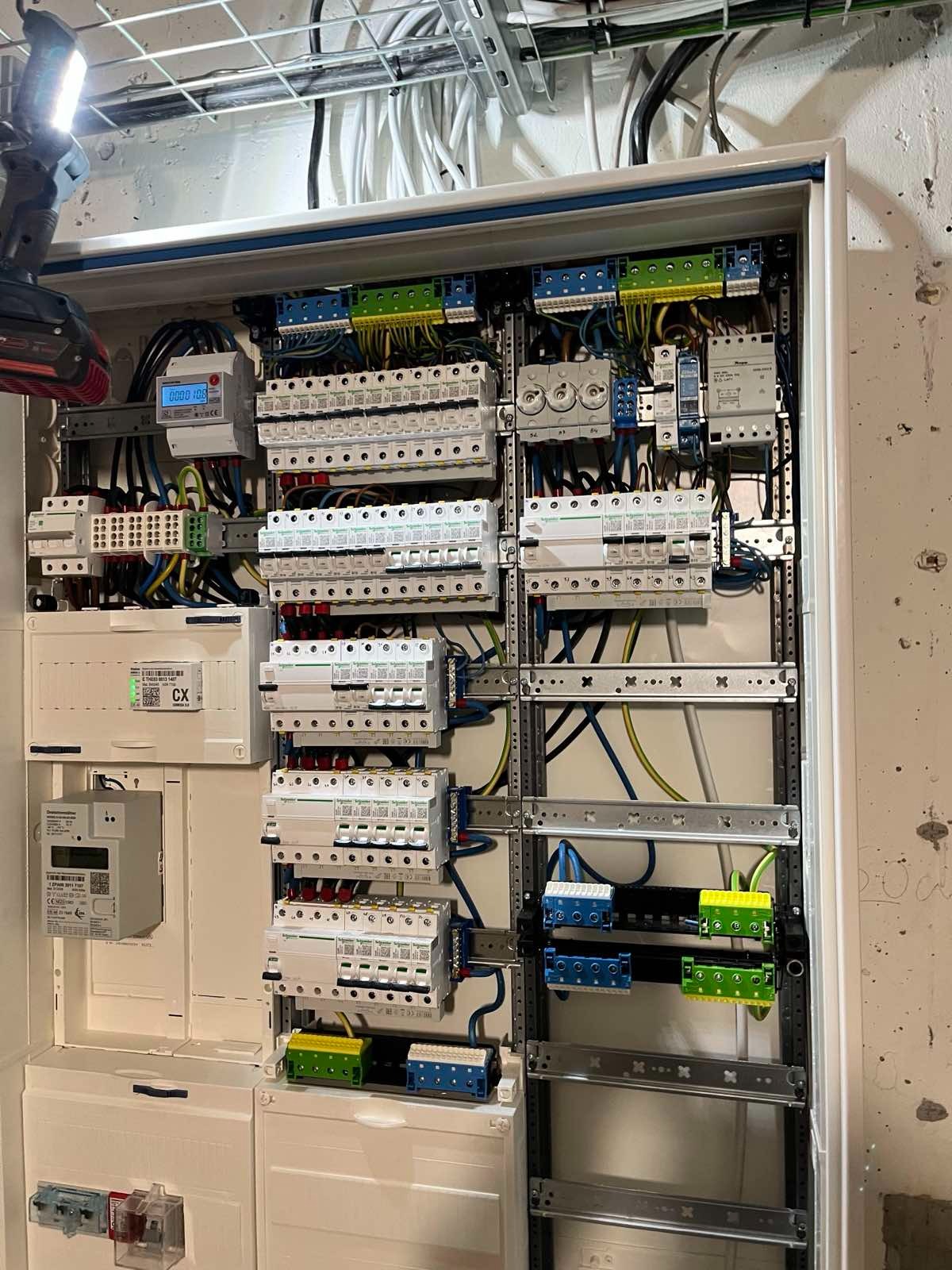 Rozdzielnica elektryczna z zamontowanymi bezpiecznikami, przewodami i licznikiem energii, oświetlona lampą roboczą. Widoczne kable w korytkach na suficie.