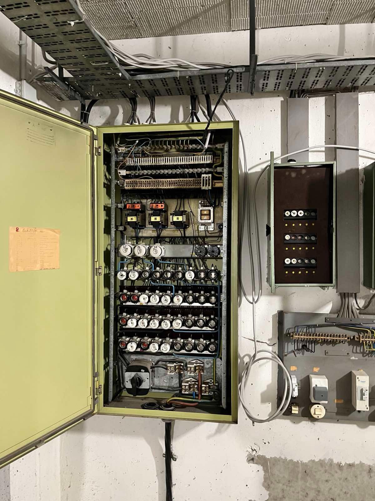 Otwarta, metalowa szafa sterownicza z licznymi bezpiecznikami i przewodami w pomieszczeniu technicznym. Dodatkowe skrzynki i kable na ścianie.