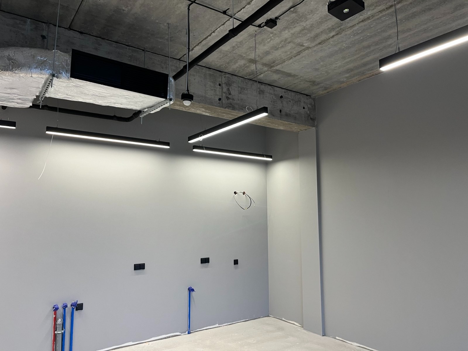 Wnętrze biurowe z surowym betonowym sufitem i liniowym oświetleniem LED. Widoczne instalacje wentylacyjne i przygotowane punkty elektryczne na ścianie.
