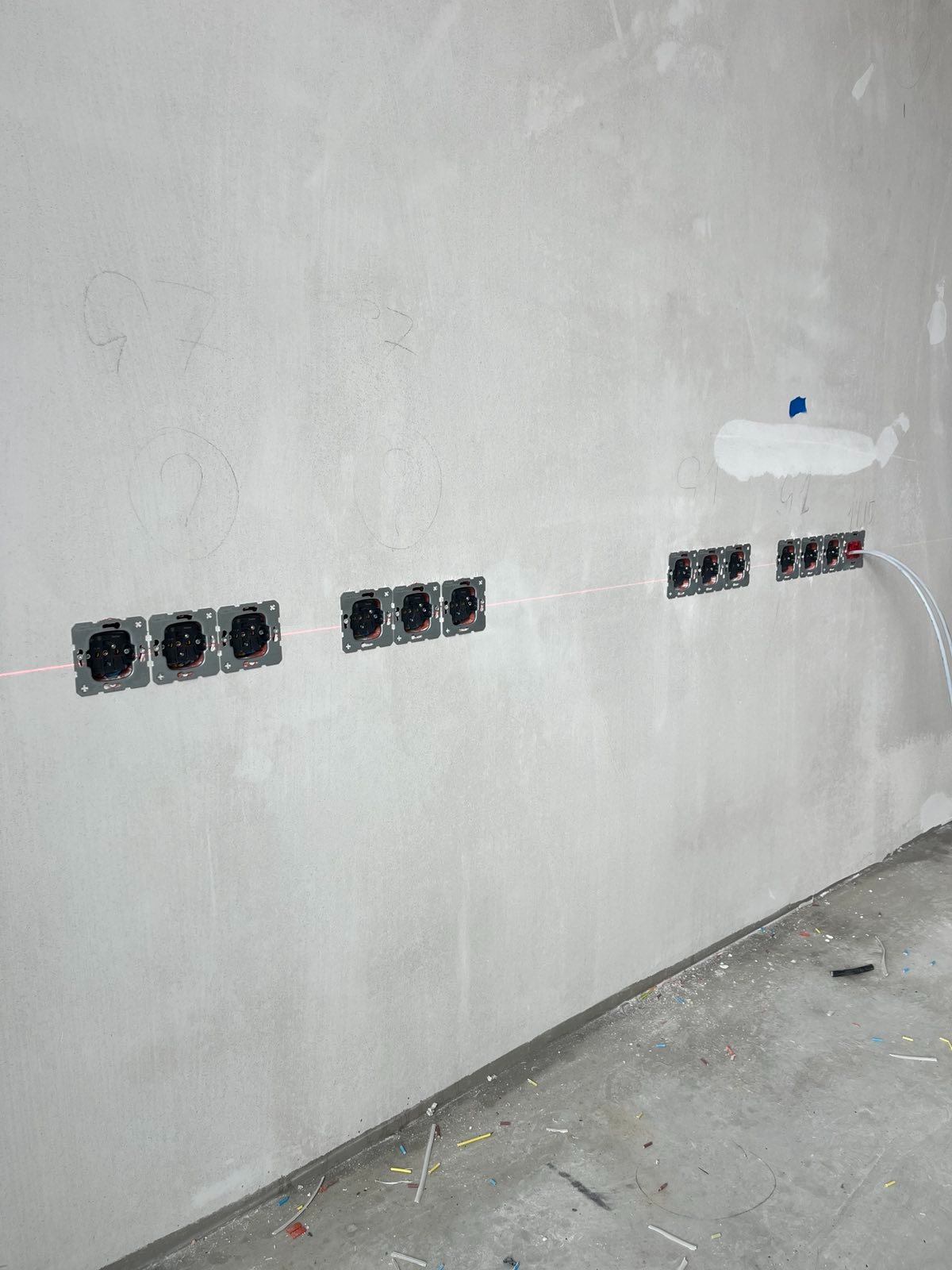 Szereg gniazdek elektrycznych w szarym murze, wyrównanych laserowo, z widocznymi przewodami. Ściana surowa, w trakcie prac wykończeniowych.