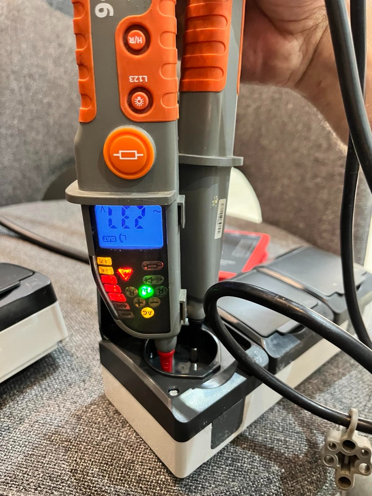 Miernik napięcia w gnieździe, wyświetlający wartość 231V, z widocznymi przyciskami i diodami LED, na tle szarej sofy i kabli.