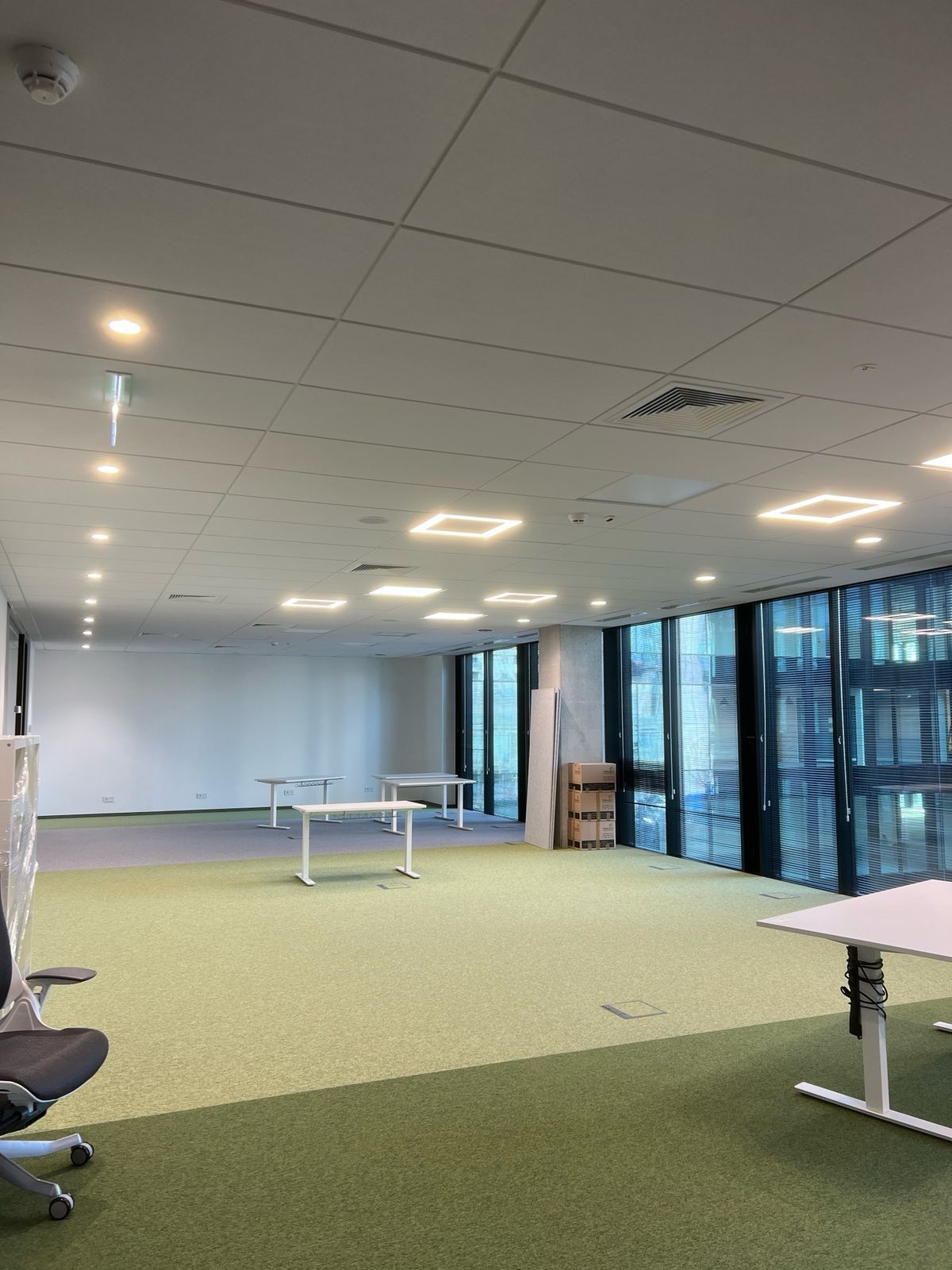 Nowoczesne biuro z kwadratowymi lampami sufitowymi, jasną wykładziną i białymi biurkami. Widoczne okna z żaluzjami i elementy wentylacji.