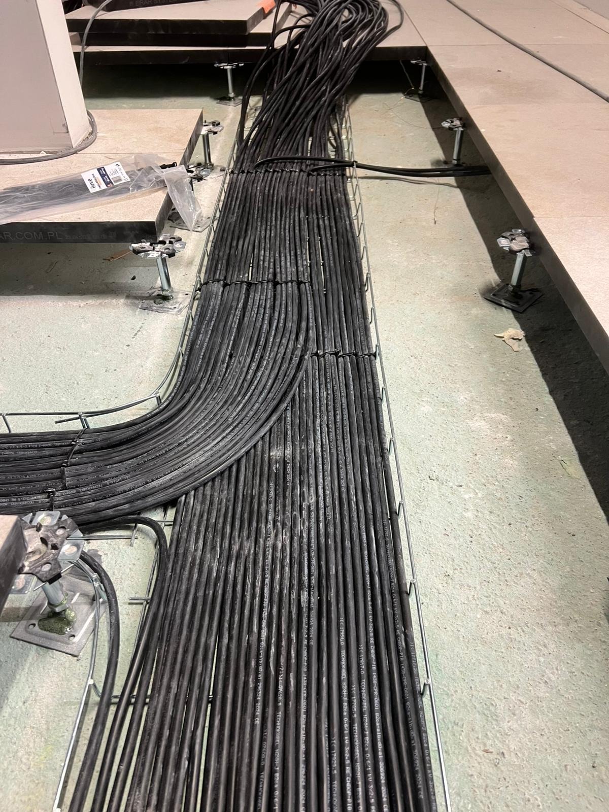 Uporządkowane kable w korycie kablowym na podwyższonej podłodze technicznej, widoczne uchwyty mocujące i fragment opakowania kabli.