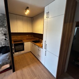 ArtMeb - Nowoczesna, kompaktowa kuchnia z białymi frontami i drewnianym blatem, zintegrowana z przestrzenią mieszkalną, widoczna z perspektywy podłogi. Minimalistyczne uchwyty.