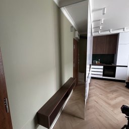 ArtMeb - Nowoczesny przedpokój z lustrem na ścianie, podwieszaną półką z ciemnego drewna i widoczną kuchnią w tle. Laserowy poziom wskazuje na precyzję montażu.