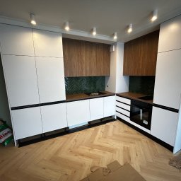 ArtMeb - Nowoczesna kuchnia z białymi frontami i drewnianymi szafkami wiszącymi, zielona mozaika na ścianie, oświetlenie punktowe, parkiet w jodełkę.