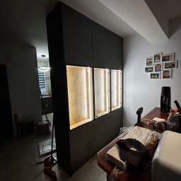 ArtMeb - Ciemna szafa wnękowa z podświetlanymi półkami z drewna, widoczna z perspektywy wnętrza, z elementami montażowymi na blacie. Zdjęcia rodzinne na ścianie.