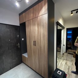 ArtMeb - Nowoczesna szafa w przedpokoju z drewnianymi frontami i czarnymi obramowaniami, z otwartą półką i siedziskiem, obok drzwi wejściowych i skrzynki narzędziowej.