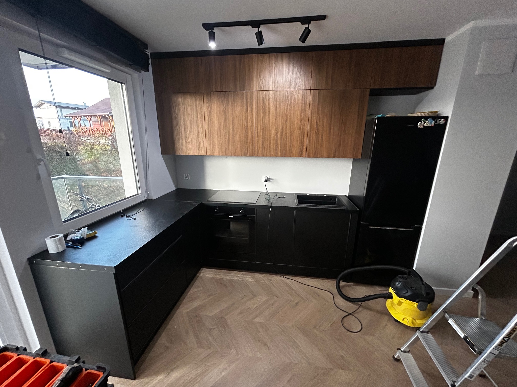 Nowoczesna kuchnia z czarnymi szafkami i drewnianymi górnymi szafkami, oświetlona reflektorami. Na podłodze parkiet w jodełkę. Widoczny odkurzacz i drabina.