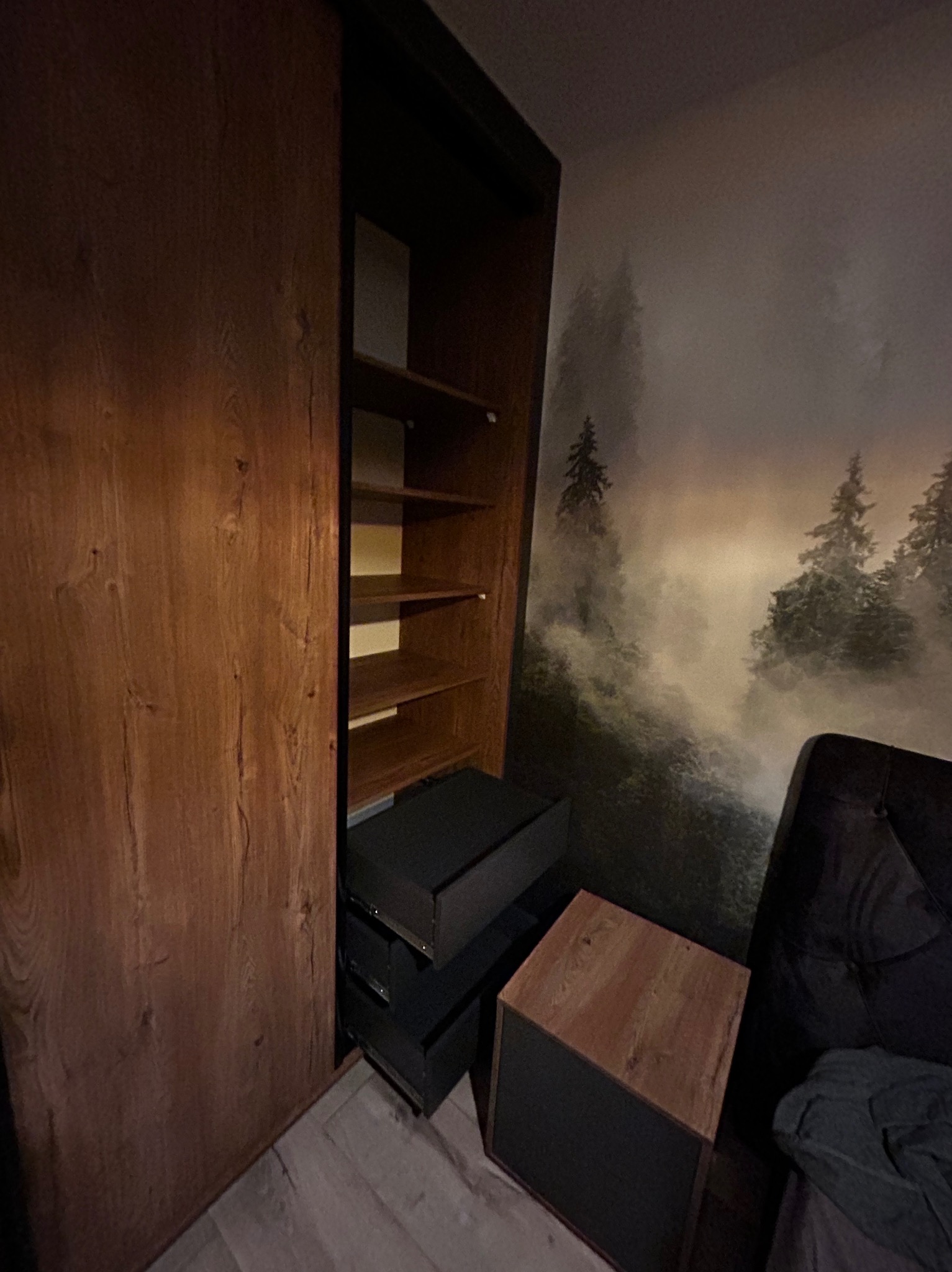 Drewniana szafa z przesuwnymi drzwiami, częściowo otwarta, ukazująca półki i szuflady. W tle tapeta z motywem leśnym i fragment sofy.