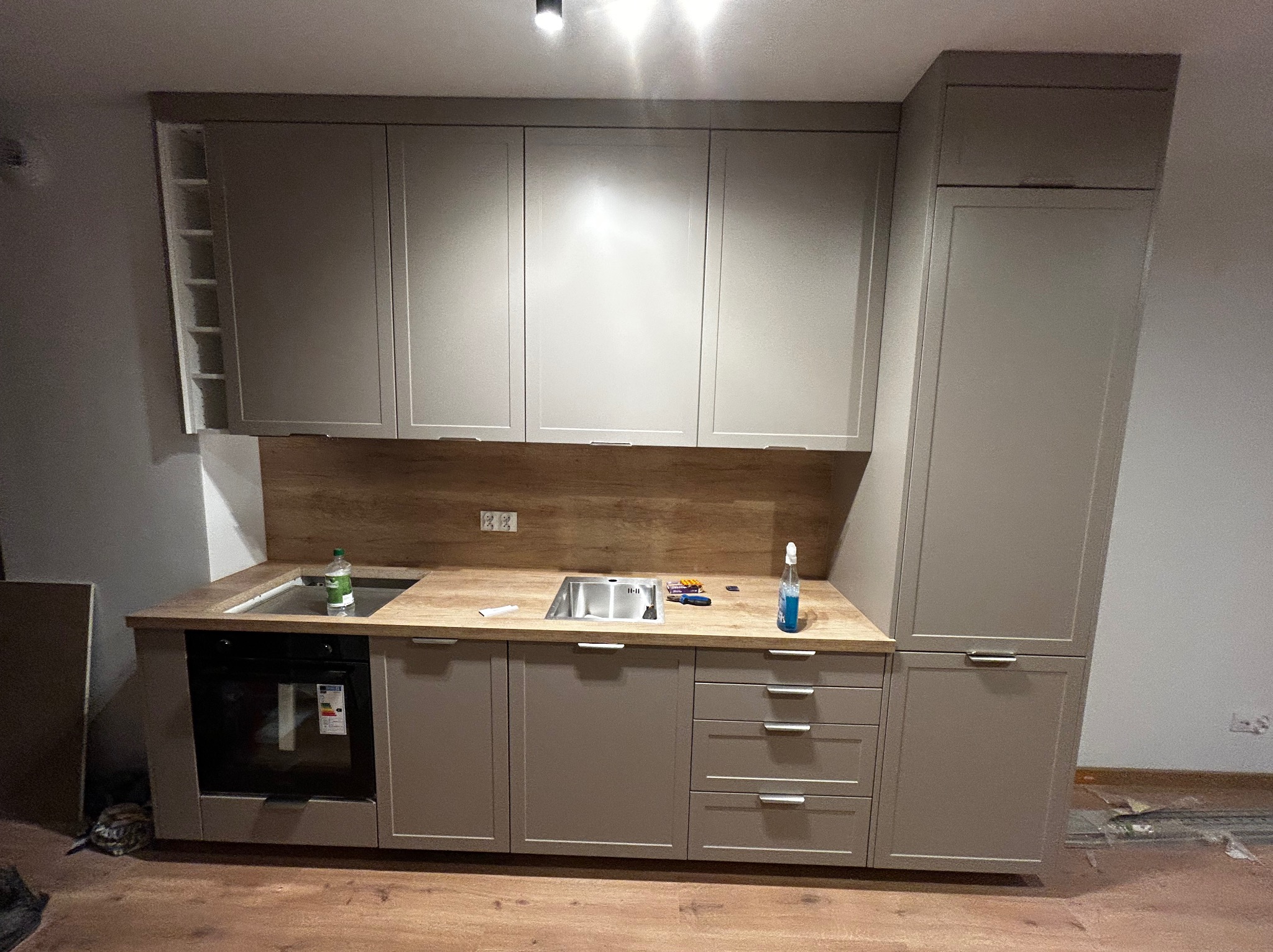 Kompletna zabudowa kuchenna w odcieniach beżu z drewnianym blatem i płytkami. Widoczne szafki górne i dolne, piekarnik oraz zlewozmywak. Minimalistyczny styl.