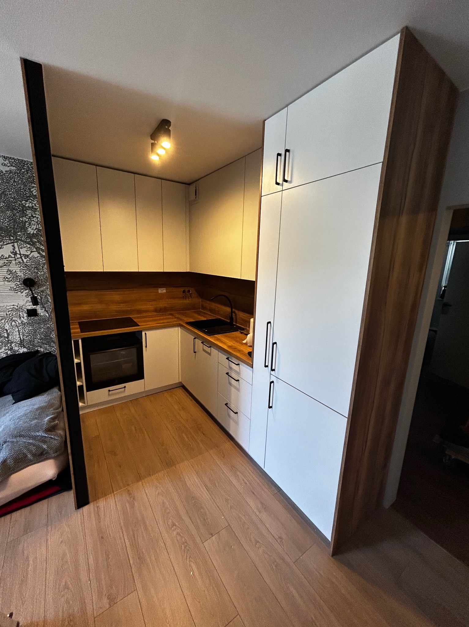 Nowoczesna, kompaktowa kuchnia z białymi frontami i drewnianym blatem, zintegrowana z przestrzenią mieszkalną, widoczna z perspektywy podłogi. Minimalistyczne uchwyty.