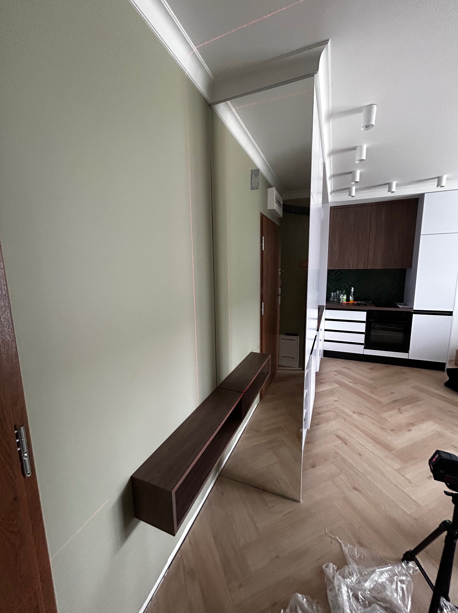 Nowoczesny przedpokój z lustrem na ścianie, podwieszaną półką z ciemnego drewna i widoczną kuchnią w tle. Laserowy poziom wskazuje na precyzję montażu.