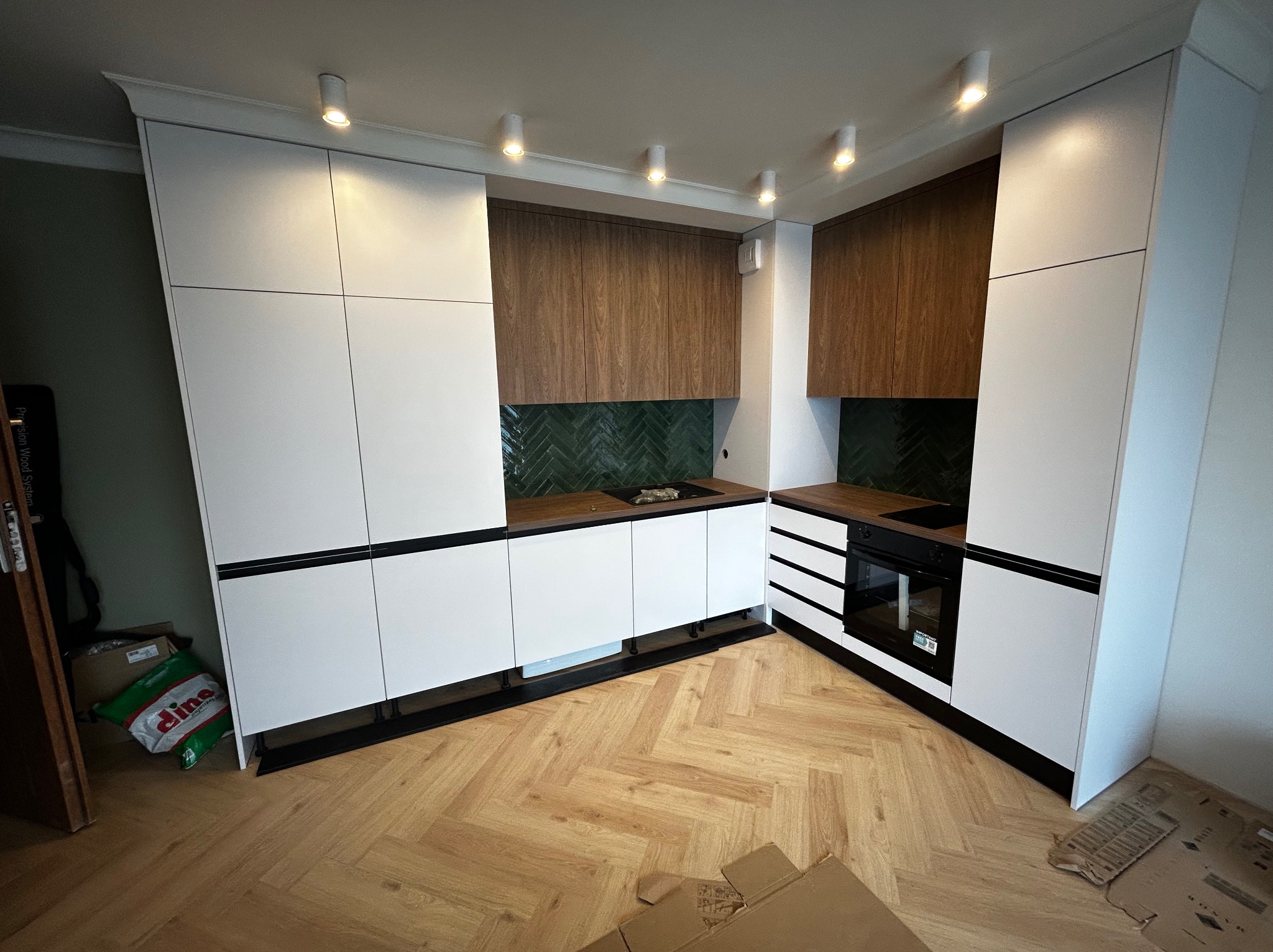 Nowoczesna kuchnia z białymi frontami i drewnianymi szafkami wiszącymi, zielona mozaika na ścianie, oświetlenie punktowe, parkiet w jodełkę.