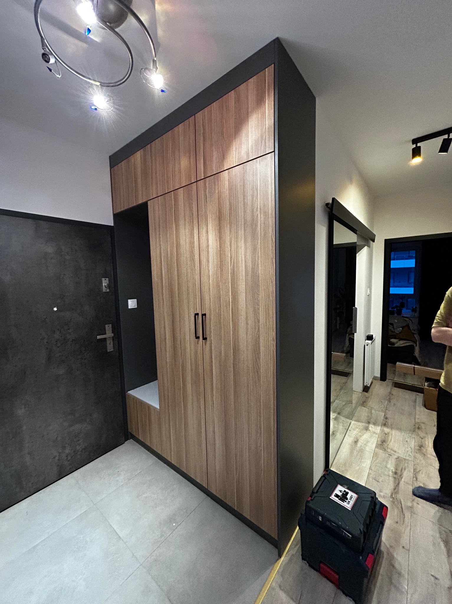 Nowoczesna szafa w przedpokoju z drewnianymi frontami i czarnymi obramowaniami, z otwartą półką i siedziskiem, obok drzwi wejściowych i skrzynki narzędziowej.