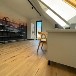 LM Wykończenia Wnętrz - Jasna drewniana podłoga w pokoju na poddaszu z oknem dachowym, biurkiem i regałem na ścianie. Widoczne naturalne usłojenie drewna i minimalistyczny design wnętrza.