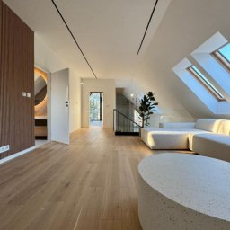LM Wykończenia Wnętrz - Jasne drewniane podłogi w przestronnym, nowoczesnym wnętrzu z oknami dachowymi i minimalistycznym wystrojem. Widoczny fragment sofy i stolika kawowego.