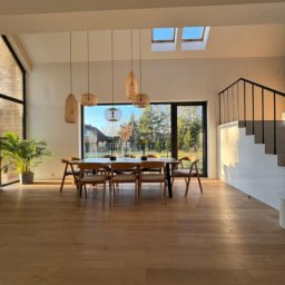 LM Wykończenia Wnętrz - Jasne, przestronne wnętrze z drewnianą podłogą, stołem jadalnym i schodami. Naturalne światło wpada przez okna dachowe i duże okno z widokiem na zieleń.